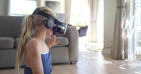 Young girl wearing vr headset exploring virtual world near couch in sunlit living room