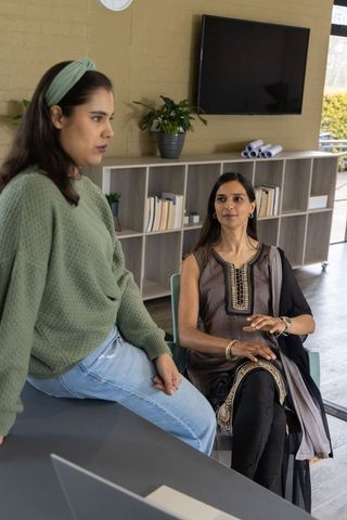 Female coworkers collaborating while sitting on desk edge in modern office workspace