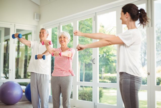 Senior couple engaging in a home workout session with a female personal trainer. They are using dumbbells and performing exercises in a bright, spacious room with large windows. This image is ideal for promoting senior fitness programs, home workout routines, and healthy lifestyle choices for the elderly.