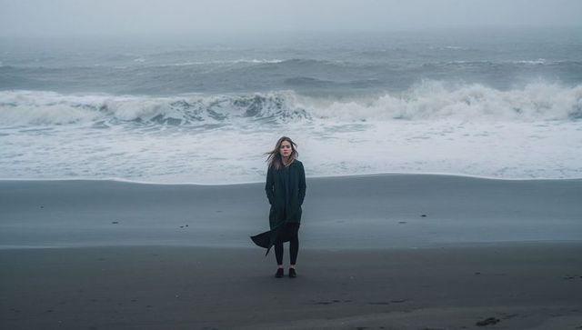 Standing solitary woman on misty dark-sand beach with windblown coat and foaming ocean