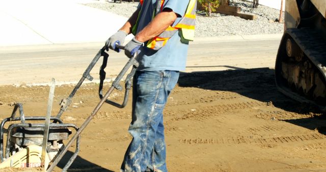 Construction worker operating a compactor machine on an unfinished road wearing safety gear including vest and gloves. Suitable for illustrating construction work, road maintenance, safety practices, or labor-related themes.