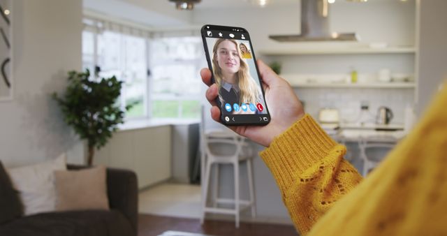 Person holding smartphone having video call in cozy home kitchen. Ideal for illustrating virtual communication, remote work, and modern lifestyle themes. Perfect for articles about digital technology, staying connected, and home office setups.