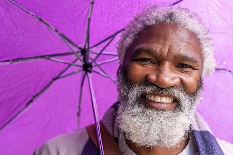 Smiling senior african american man holding purple umbrella in rain, vibrant portrait