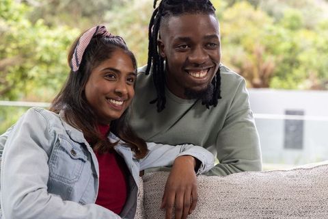 Smiling diverse couple relaxing on cozy sofa near glass railing with terrace greenery