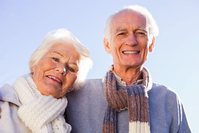 This warm and cheerful image depicts an elderly couple embracing outdoors on a sunny day, both wearing winter clothing including scarves. Perfect for use in advertisements or articles related to senior care, relationships in later life, health and wellness for the elderly, and lifestyle content aimed at senior citizens.
