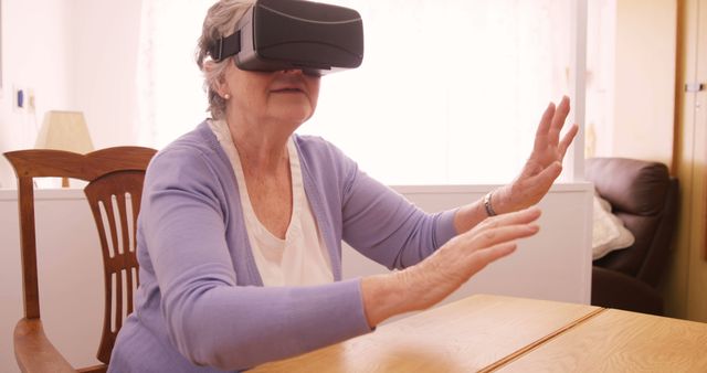 Elderly woman actively engaging with a virtual reality headset while seated in a modern living room. Ideal for showcasing technology adoption among seniors, innovative ways the elderly can stay entertained, and the positive impact of VR in social and recreational activities. Useful in articles, brochures, and websites focused on technology, senior living, and modern health care solutions.