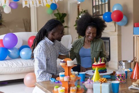 Couple celebrating birthday at home blowing candles on rainbow cake with balloons