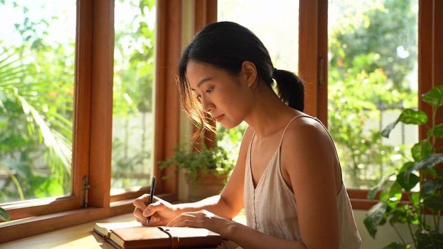 Journaling at sunlit window with indoor plants, woman writing in leather notebook, calm morning