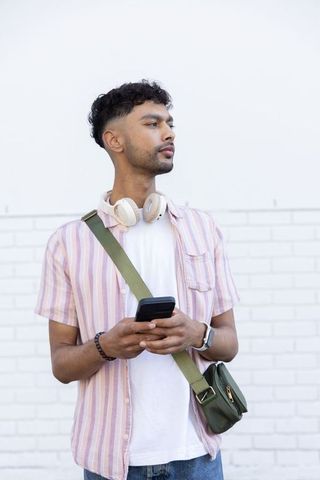 Indian young man wearing headphones holding smartphone against white brick wall casual