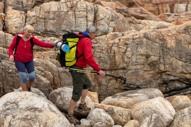 Senior Caucasian couple enjoying an adventurous hike on rocky terrain. Both are equipped with backpacks and hiking poles, showcasing their active lifestyle and love for nature. Ideal for promoting outdoor activities, senior fitness, adventure tourism, and retirement lifestyle.