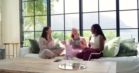 Multicultural friends laughing and drinking coffee on sunlit couch in casual loungewear