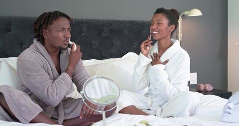 African american couple enjoying cozy skincare routine with handheld facial devices on bed