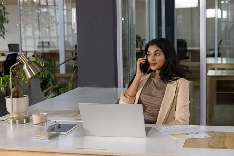 Indian professional in beige blazer talking on phone at modern glass office desk