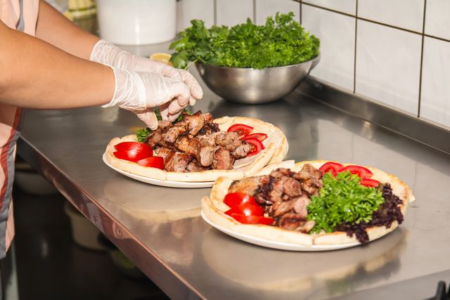 Chef preparing shawarma sandwiches in a commercial kitchen. The chef is adding grilled meat to sandwiches with gloves, ensuring proper hygiene. Sandwiches are served on pita bread with fresh tomatoes, parsley, and herbs. Suitable for presentations on culinary arts, food hygiene practices, and restaurant marketing.