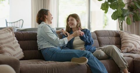 Two women laughing and holding hands on cozy tufted sofa in sunlit living room