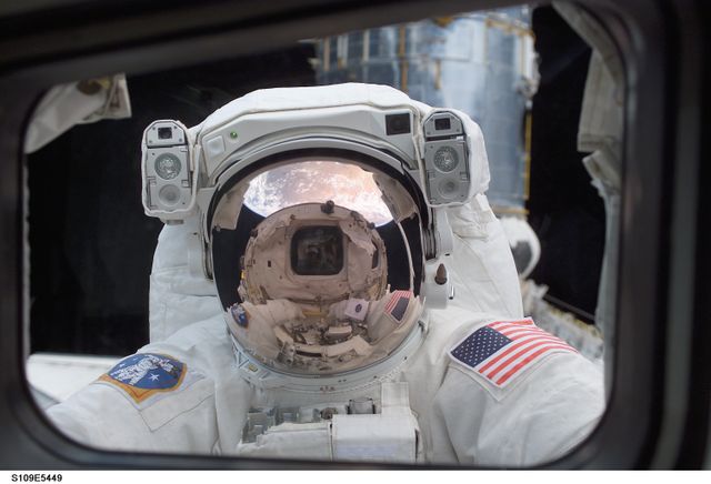 Astronaut engages in spacewalk, shown peering into Shuttle Columbia's crew cabin. Reflection on helmet visor showcases Earth's hemisphere and shuttle, creating a distinctive view. Suitable for topics on space exploration, NASA missions, spacewalks, and the Hubble Space Telescope.