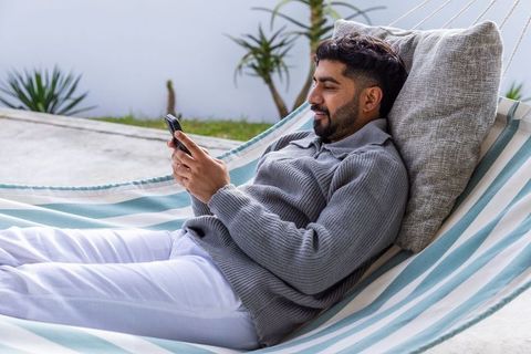 Asian man relaxing in striped hammock on sunny patio texting on smartphone near succulents
