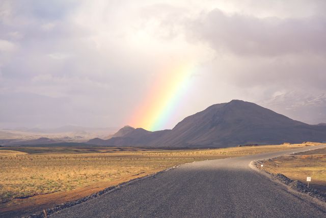 Winding road leads to a distant rainbow over serene mountains. Scenic view showcases natural beauty. Ideal for travel brochures, inspirational posters, or desktop wallpapers.