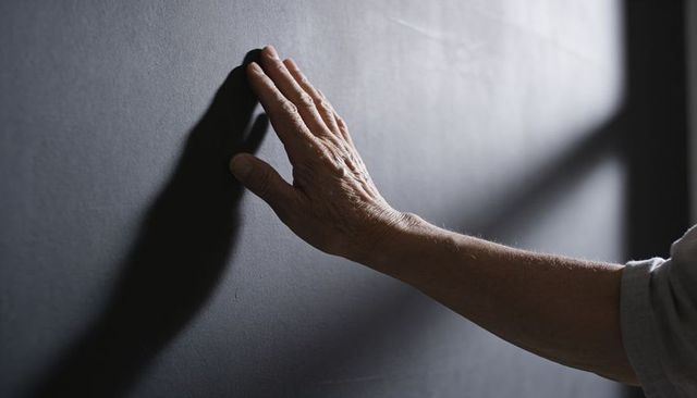 Senior hand pressing against dark wall casting diagonal window shadow, forearm wrinkles
