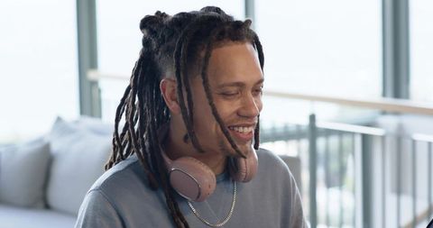 Smiling african american man wearing pink headphones and chain, sitting in sunlit modern loft