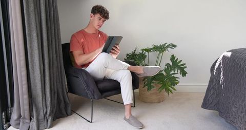 Young man reading tablet in modern bedroom chair with houseplant and natural light