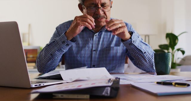 Senior man wearing glasses engaged in work at home office, reviewing documents and using a laptop. Ideal for themes related to remote work, seniors in business, home office setups, and professional concentration.