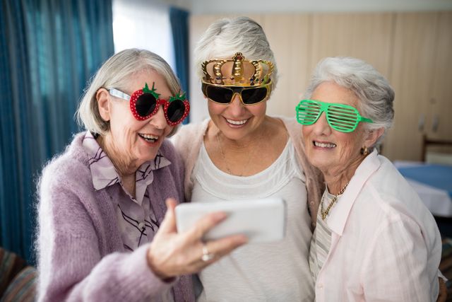 Three senior women wearing fun novelty glasses are taking a selfie with a smartphone in a nursing home. They are smiling and appear to be enjoying their time together. This image can be used to depict themes of friendship, joy, and the positive aspects of aging. It is suitable for use in advertisements, articles, or social media posts related to elderly care, retirement communities, and senior lifestyle.