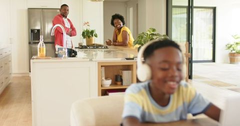 African american family relaxing in sunlit kitchen with child in headphones using tablet