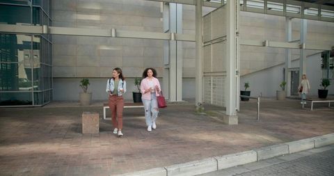 Two women walking in modern plaza carrying coffee and tote bag casual urban lifestyle