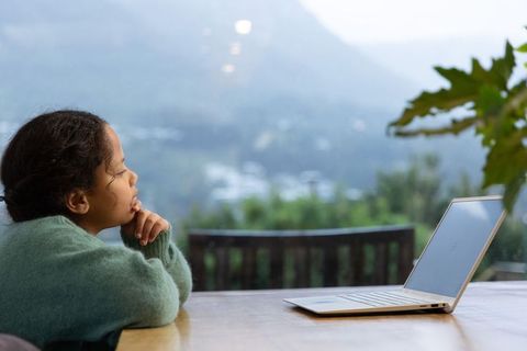 Young girl gazing through window at misty mountain valley during remote learning with laptop on tabl