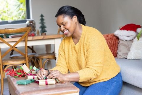 African american woman wrapping holiday gifts at home in cozy mustard sweater