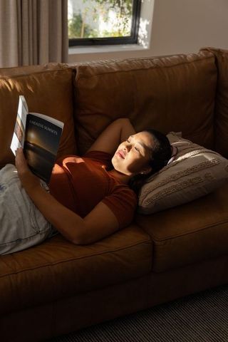 Mid adult asian woman reclining on leather sofa reading 'endless sunsets' in warm sun