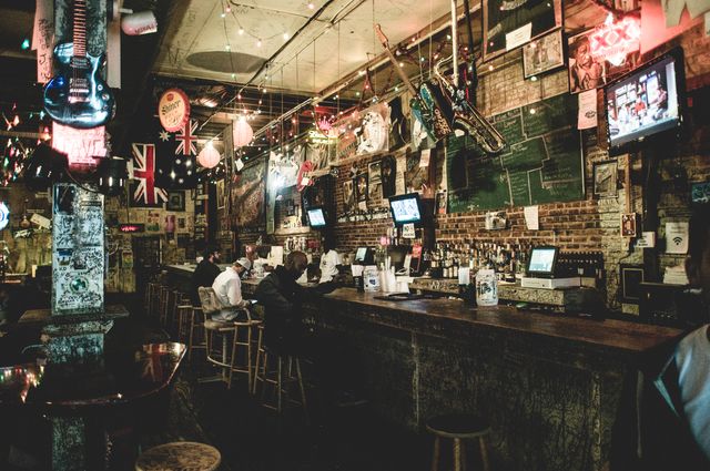 Rustic bar with a lively atmosphere, featuring hanging musical instruments, wall decorations, and multiple televisions. Patrons sit on stools by the bar, enjoying drinks and conversation. Perfect for illustrating nightlife, social scenes, or entertainment venues.