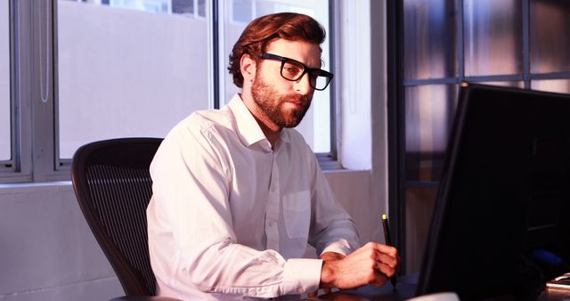 Young professional man in white shirt and glasses working at desk with computer in a modern office environment. Useful in business, technology, or corporate context highlighting focused work, office environment, and workplace productivity.