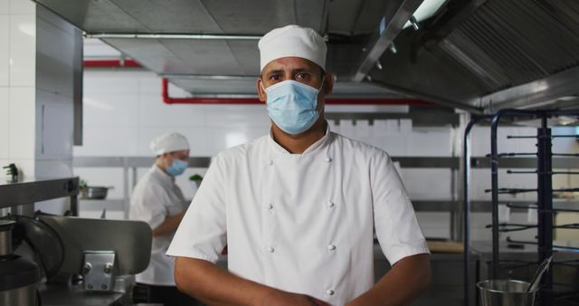 Portrait of biracial male chef wearing face mask with arms crossed. Health and hygiene in restaurant kitchen during coronavirus covid 19 pandemic.