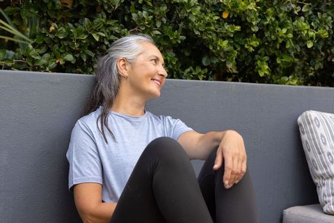 Senior woman relaxing on outdoor bench wearing gray tee and black leggings, green hedge
