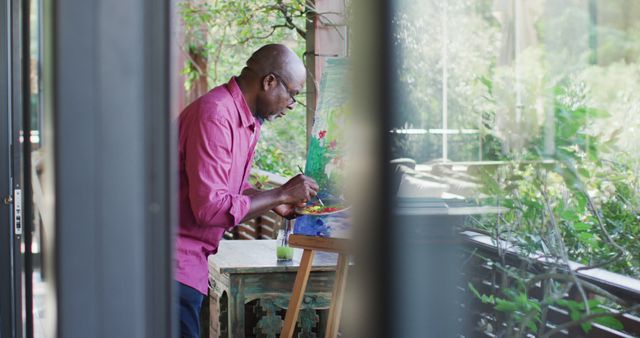African American man engrossed in painting, standing in an art studio that blends with lush greenery and natural light. Ideal for illustrating artistic creativity, serenity in nature, and the focus of an artist at work. Suitable for articles about art therapy, personal expression, creative hobbies, or the benefits of a natural setting for creativity.