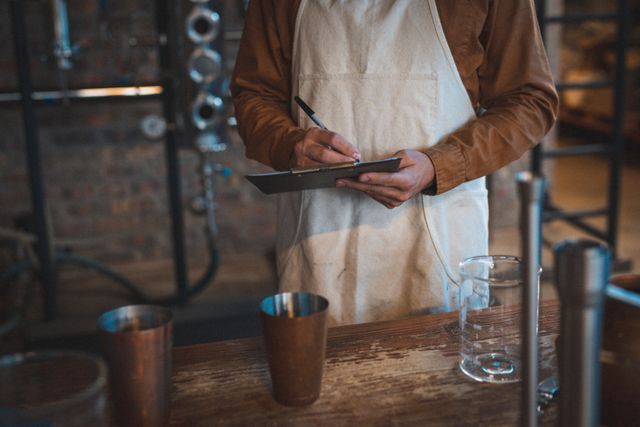 Craft gin distiller taking notes on clipboard in distillery. Ideal for content about small businesses, craft alcohol production, artisan manufacturing, and industrial processes. Useful for articles, blogs, and marketing materials related to the craft beverage industry.