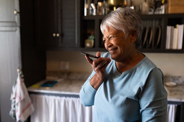 Senior African American woman standing in kitchen, smiling while talking on smartphone. Ideal for use in articles or advertisements about retirement lifestyles, technology use among seniors, communication during self-isolation, and the impact of the COVID-19 pandemic on daily life.