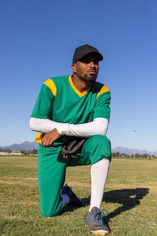 African american baseball player kneeling on field wearing green uniform and black cap