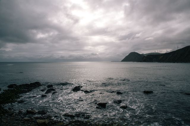 A dramatic scene with dark overcast clouds covering the sky and a calm sea reflecting the subdued light. The rocky shoreline adds texture and contrast to the picturesque coastal view. Perfect for use in projects related to nature, seascapes, tranquility, atmospheric landscapes, or any creative endeavor needing a moody, serene background.
