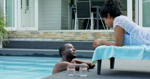 Black man and indian woman relaxing and chatting at pool edge on backyard deck