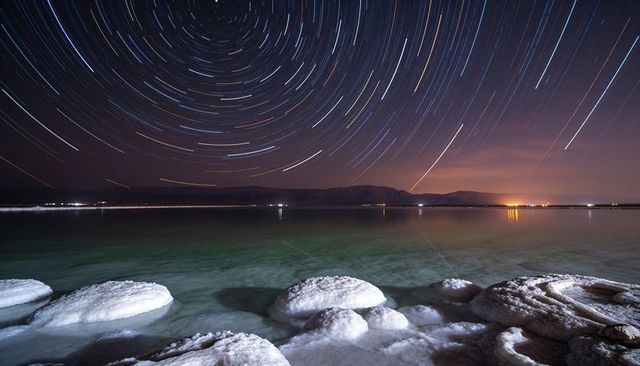 Salt-crusted shoreline with circular star trails sweeping night sky over tranquil saline waters