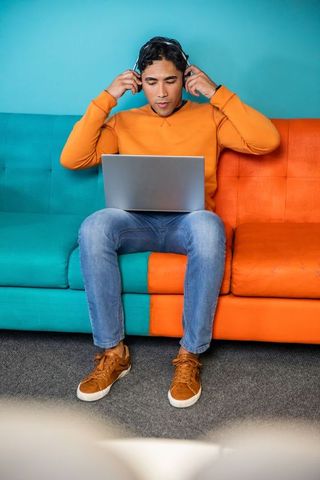 Indian man working on laptop while adjusting headphones on vibrant two-tone sofa in modern workspace