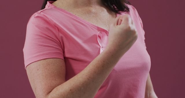 Mid section of a woman with pink ribbon on her chest clenching her fist against pink background. breast cancer awareness concept
