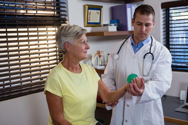 Physiotherapist assisting senior woman with stress ball exercise in clinic. Ideal for use in healthcare, rehabilitation, and elderly care promotions. Useful for illustrating physical therapy sessions, patient care, and wellness programs for seniors.