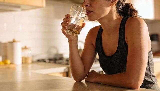 Sunlit woman drinking lemon water while leaning on kitchen counter in athletic tank top
