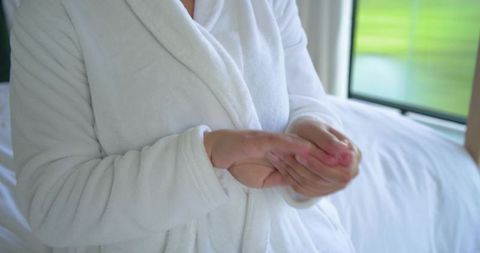 Woman in white bathrobe holding hands at chest relaxing in sunlit cozy bedroom