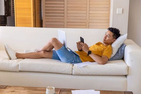 Young asian man reclining on sofa scrolling smartphone and working on laptop at home