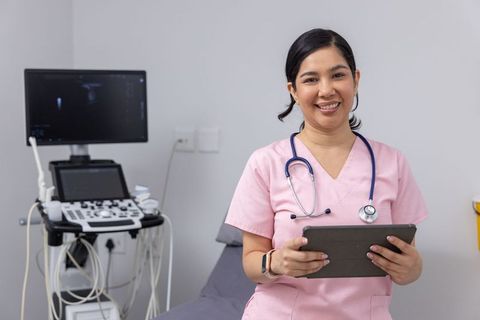 Smiling nurse in pink scrubs holding tablet next to ultrasound machine